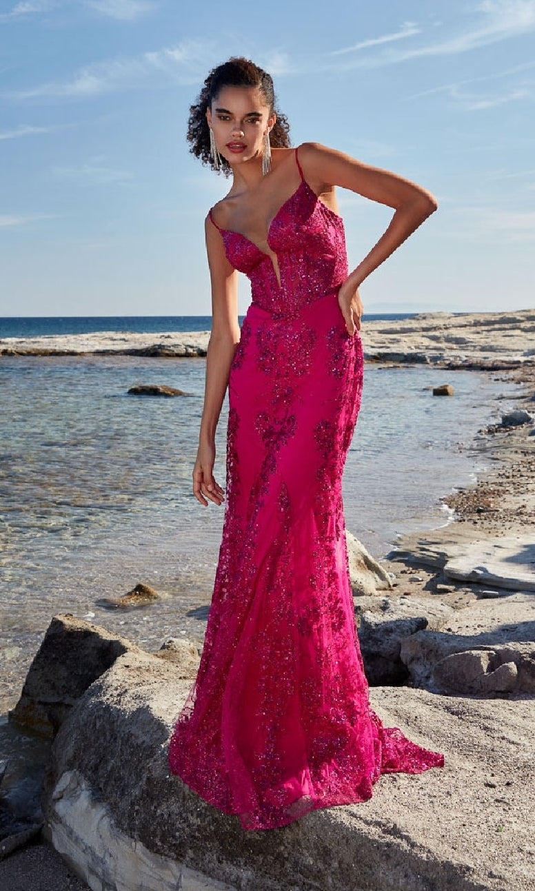A woman in the Strappy-Back Long Prom Dress: Aerie Couture 206, a vivid pink gown, stands on rocks near a calm, rocky shore beneath a clear blue sky.